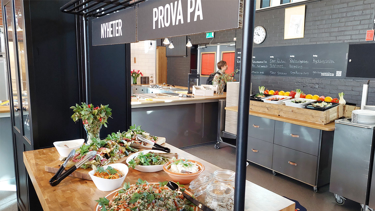 En provsmakningsstation i en skolmatsal märkt "Nyheter" och "Prova På" med skålar av sallad, wraps och grönsaker. En griffeltavla med dagens rätter och en salladsbar syns i bakgrunden.