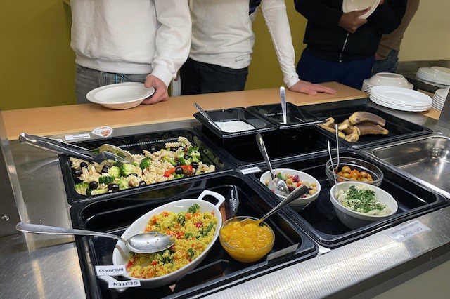 Elever står vid buffévagn i skolrestaurang, redo att ta mat