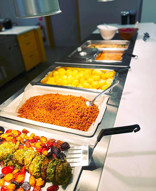 Buffé med mat för skolmåltid. I förgrunden syns örtbiffar med rostade rotfrukter och bulgur, samt kokt potatis och sås längre bak.