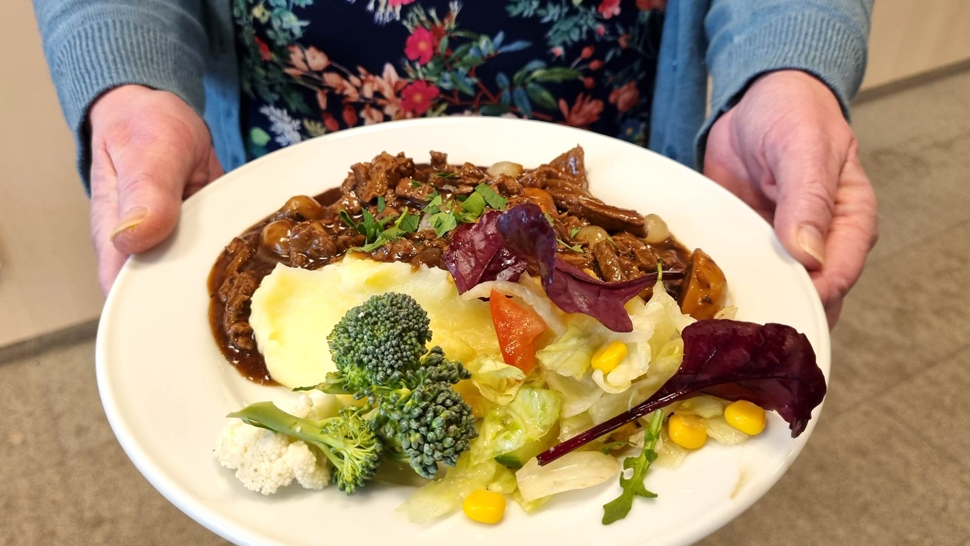 Äldre kvinna håller i tallrik med köttgryta och potatismos samt grönsaker