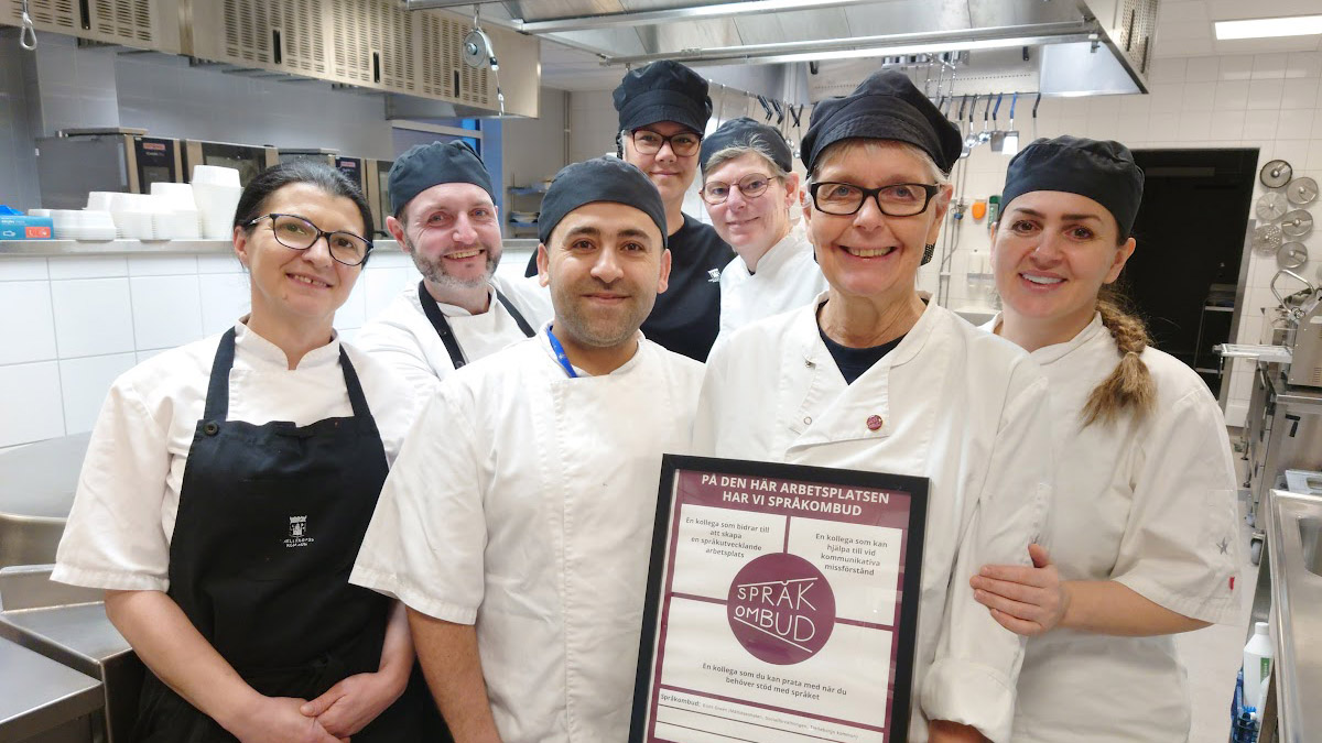 En grupp med glada kockar i en storköksmiljö. En av dem, Bodil Green, håller i ett certifikat som visar att arbetsplatsen har ett språkombud