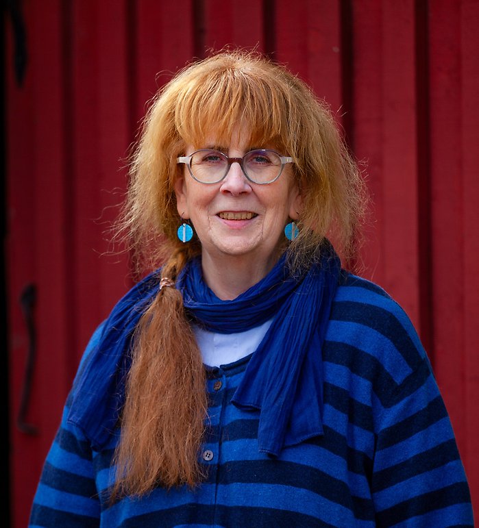 Lena har rött hår och blå kläder. Hon står framför en rödlada