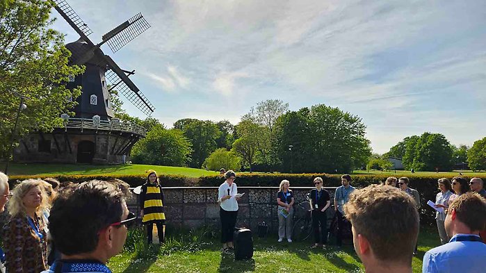 Personer utomhus i Slottsparken i Malmö. Det är en guidad tur. Guiden är iklädd en humledräkt. Åhörarna står i en halvcirkel och lyssnar.