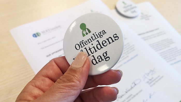 En hand som håller fram en "badge" med orden Offentliga måltidens dag.