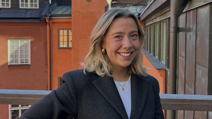 Kristelle Schepens har mörk kavaj och vit t-shirt. Hon står på en balkong med solbelysta hus i bakgrunden