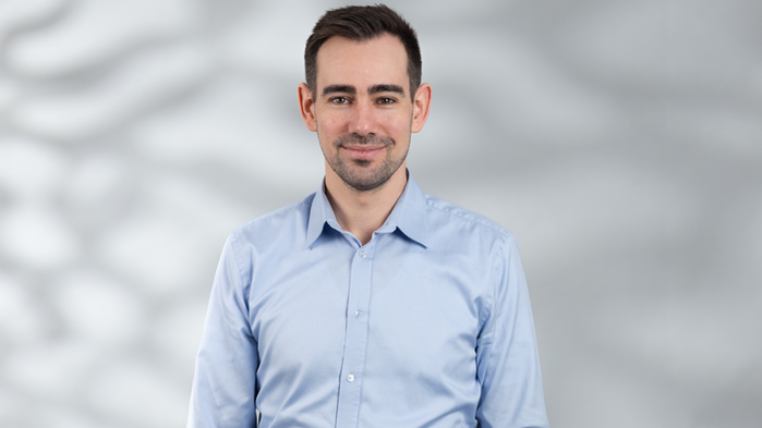 Johannes Hansson har ljusblå skjorta och mörk kort hår och står framför en vit bakgrund