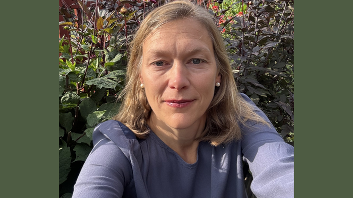 Charlotta Rydberg har lila tröja och blont hår. Hon står utomhus med blommor i bakgrunden