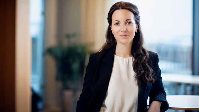 Mathilda Hollertz har mörk kavaj och vit skjorta. Hon står vid ett stort fönster i bakgrunden